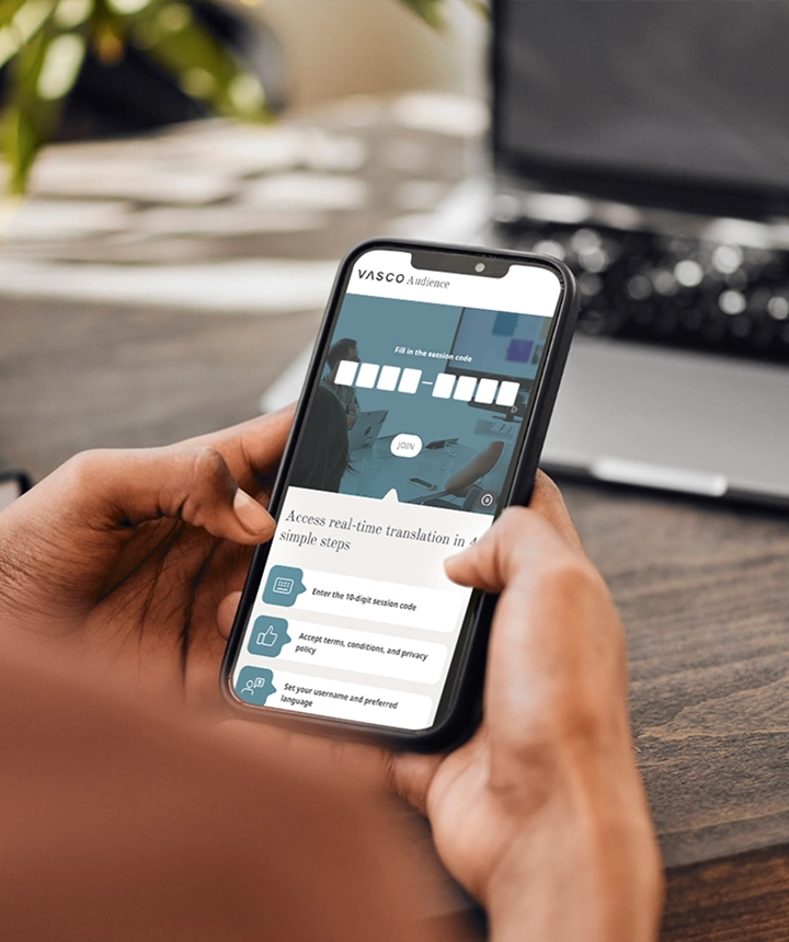 Egy személy váll felett nézve, aki egy okostelefont tart a kezében, amelyen a Vasco Audience webes felülete látható. A képernyőn egy „Írja be a munkamenet kódját” nevű beviteli mező látható egy fehér „CSATLAKOZÁS” gombbal, amely alatt egy útmutató felirat olvasható: „Valós idejű fordítás 4 egyszerű lépésben”. A háttérben egy elmosódott fa íróasztal és egy laptop látható. 