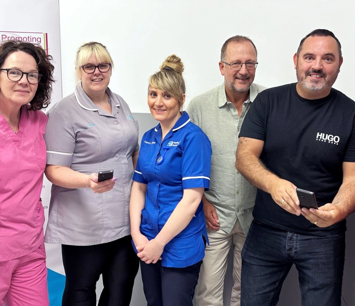 Öt mosolygós ember áll együtt egészségügyi öltözékben, ketten közülük tolmácskészüléket tartanak.