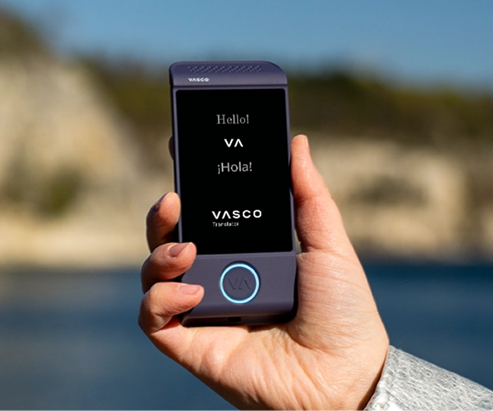Közelkép egy kézről, amely egy lila Vasco Translator M4 elektronikus fordítógépet tart a kezében, világos kültéri háttér előtt. A képernyőn a „Hello!”, „¡Hola!” üdvözlések és a „VASCO Translator” logó látható.