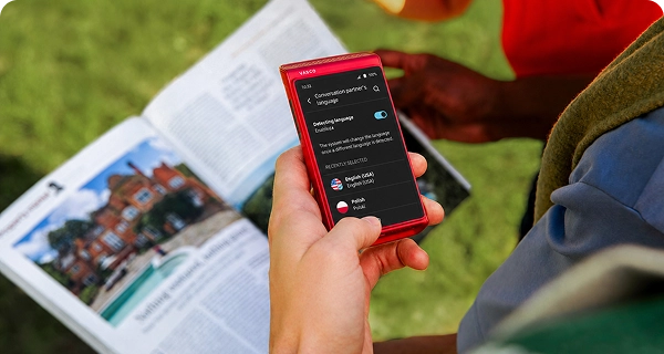 Valaki egy piros Vasco Translator Q1 készülékkel fényképet készít egy újságról. A képernyőn megjelenik a nyelvválasztó menü, amely az újságból automatikusan felismert szöveg fordításához szolgál.